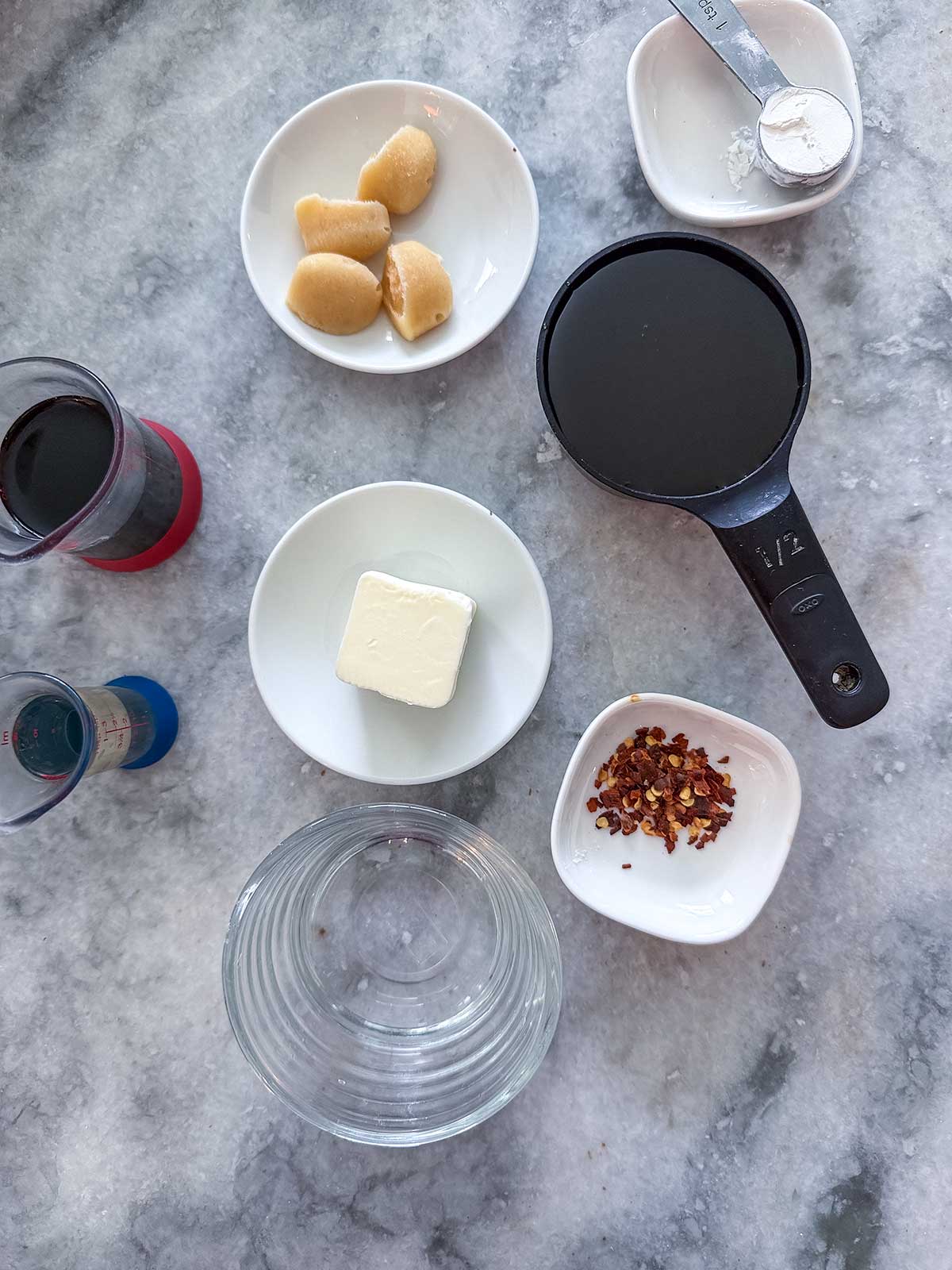 Various ingredients sit on a gray marble surface, ready to make Honey Garlic Salmon Bites in the Air Fryer, including a black measuring cup with dark liquid, a small plate of butter, brown paste, red pepper flakes, a clear glass, and two small cups with liquids.