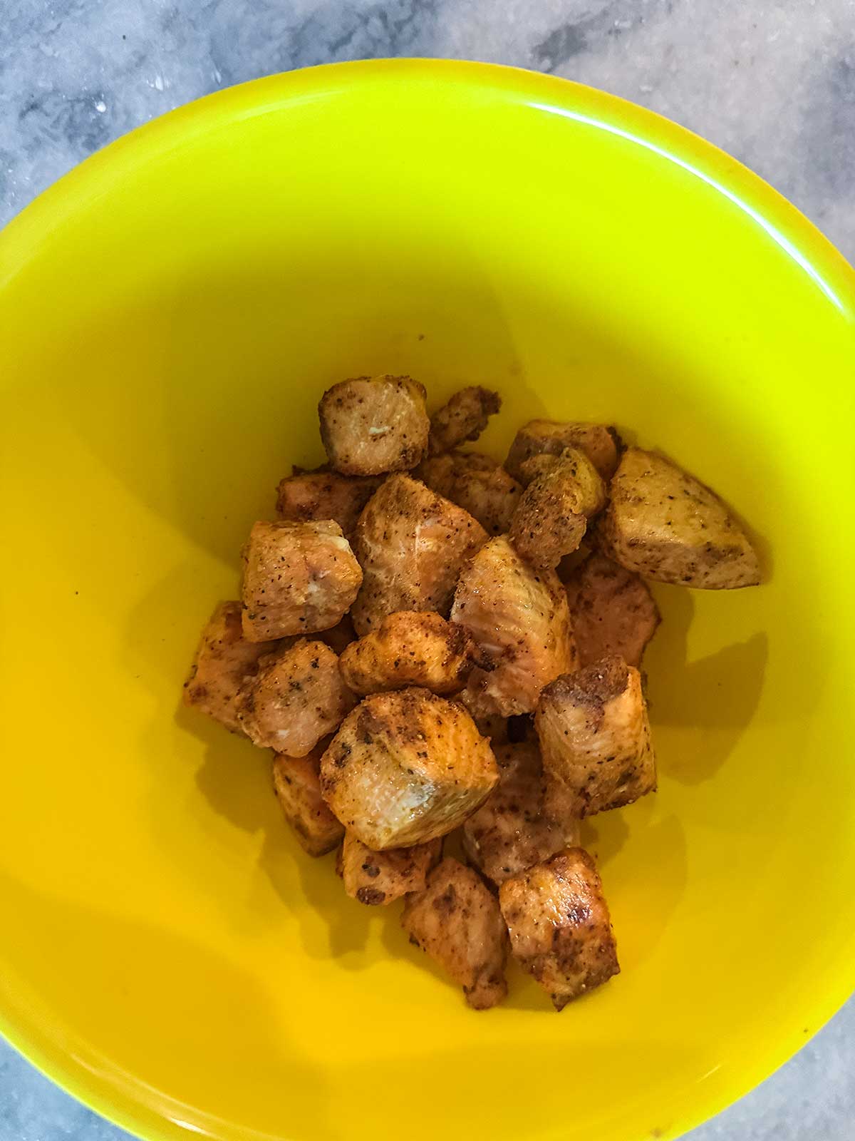 Seasoned, cooked salmon pieces in a bright yellow bowl on a gray surface. The chicken is cut into bite-sized chunks and appears to be well-spiced—perfect for pairing alongside air fryer favorites or honey garlic salmon bites.