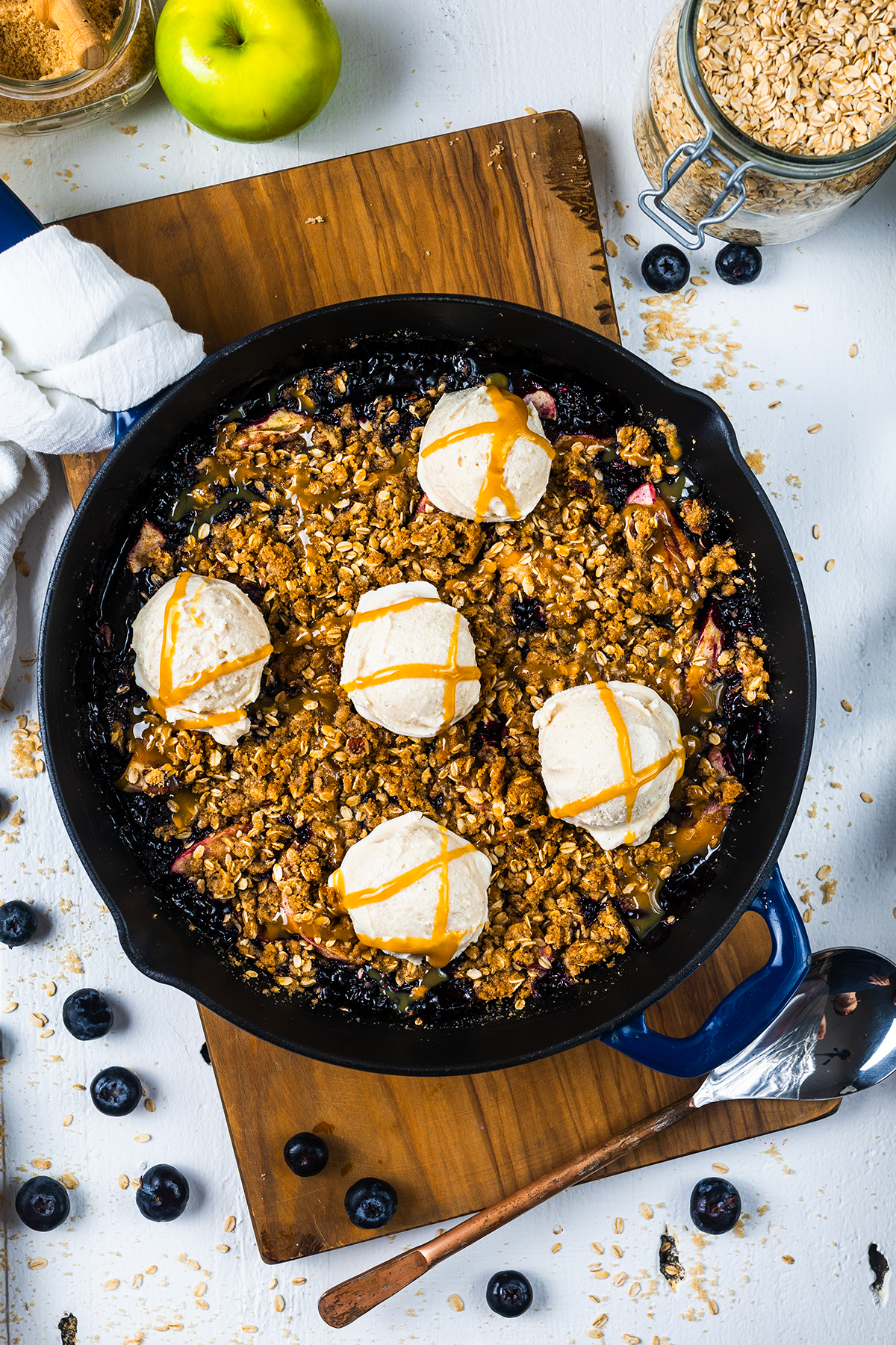 An Apple Blueberry Crisp sits on a wooden board, its warm skillet filled with juicy apples and blueberries beneath a golden oat crumble. Five scoops of vanilla ice cream slowly melt into the crisp, mingling with a decadent caramel drizzle. The scene is completed by fresh blueberries, oats, and a green apple scattered around for an inviting touch.