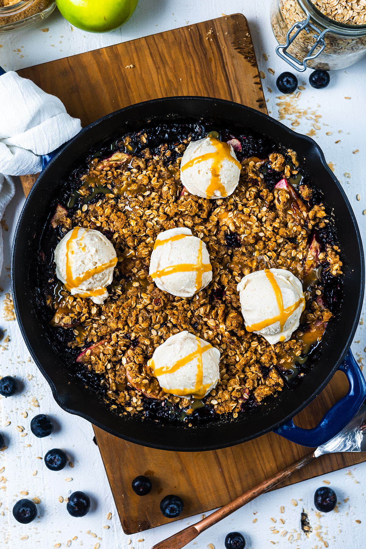 A skillet Apple Blueberry Crisp topped with six scoops of vanilla ice cream and drizzled with caramel sauce, sitting on a wooden board with fresh blueberries scattered around.