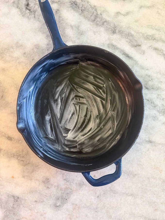 A cast iron skillet with a blue handle, perfect for baking an apple blueberry crisp, is coated with a layer of butter or oil and sits on a light-colored marble countertop.