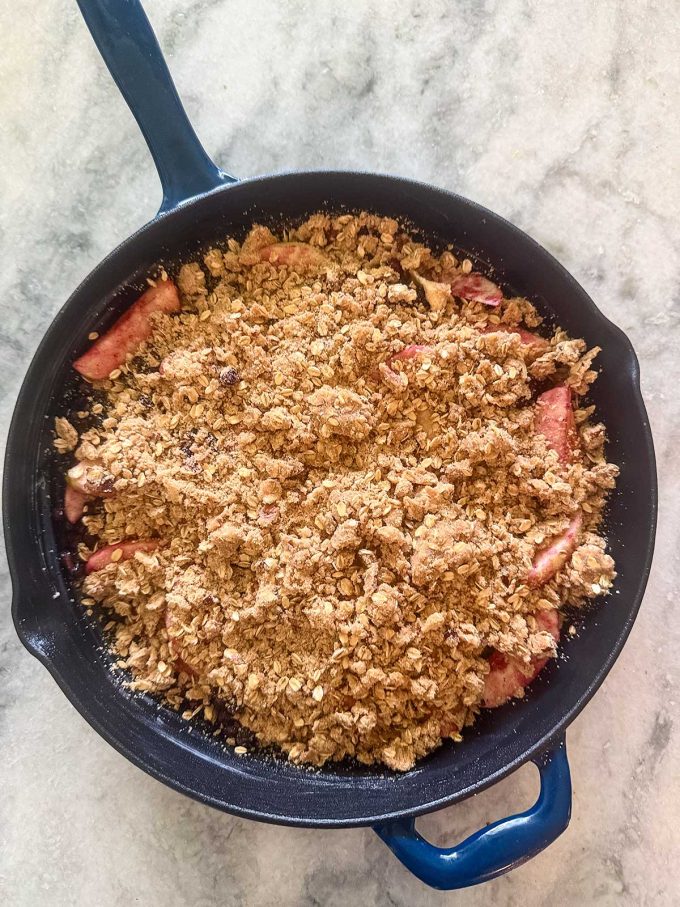 A black skillet filled with a delicious Apple Blueberry Crisp, topped with a golden brown oat and crumb mixture, sits on a light marble surface.