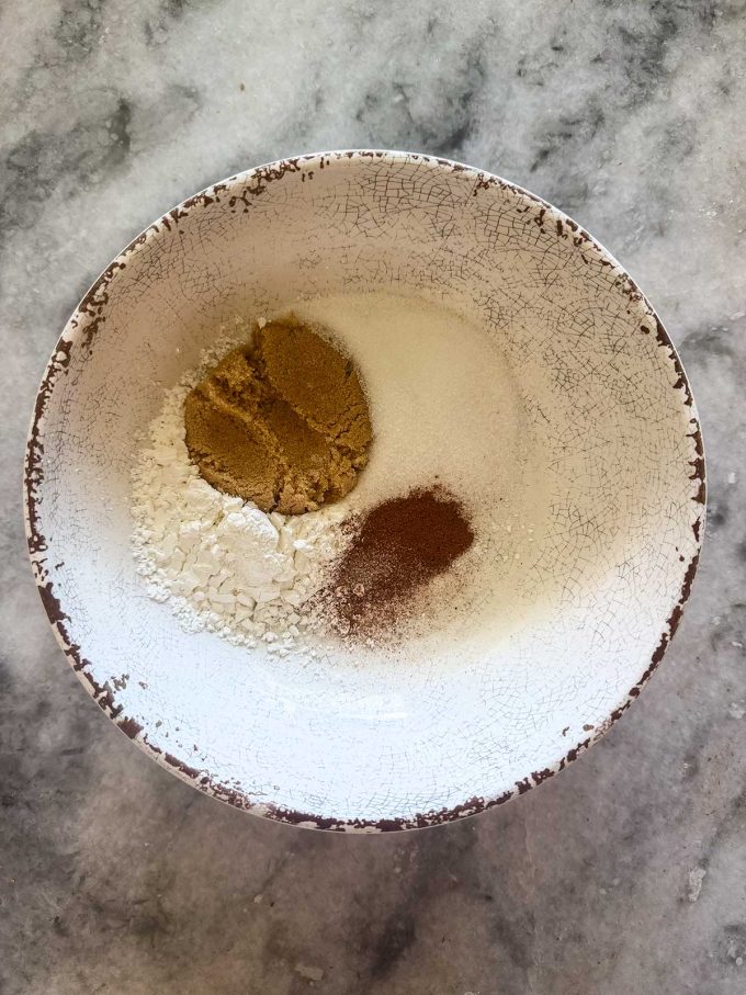 A white bowl with cracked edges, perfect for preparing an Apple Blueberry Crisp, contains white flour, granulated sugar, brown sugar, and ground cinnamon, arranged separately on a gray marbled countertop.