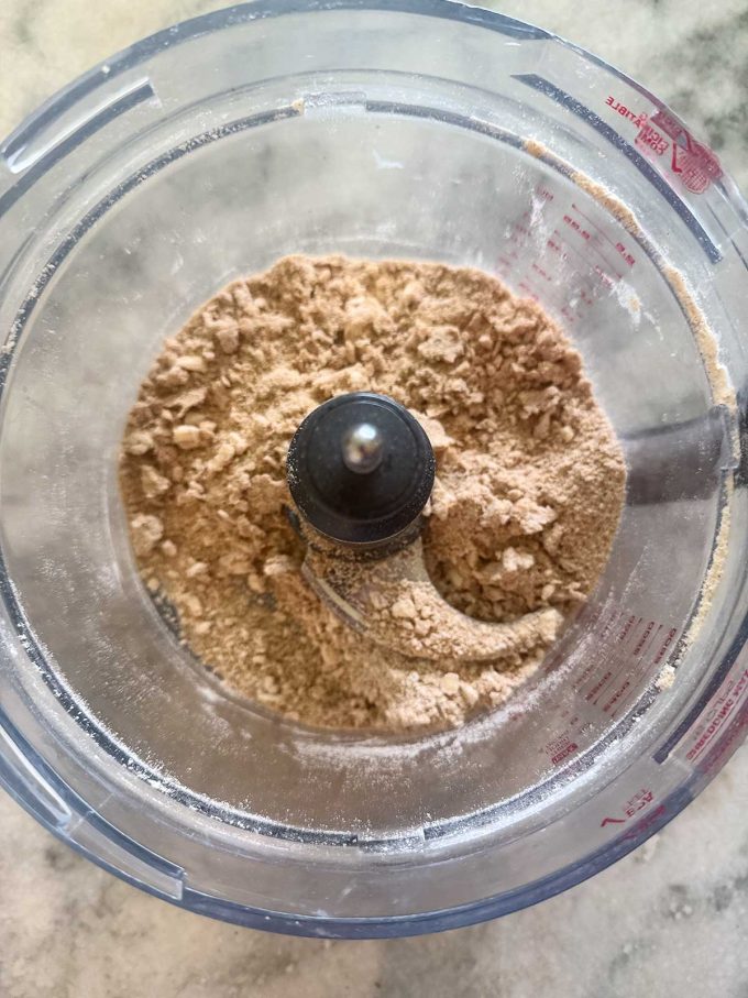 A food processor bowl contains finely ground, light brown crumbs, perfect for a blueberry crisp, on a countertop, viewed from above.
