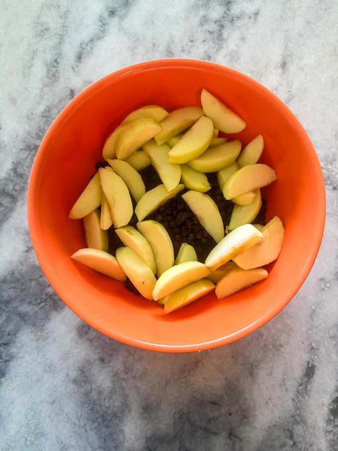 Orange bowl on a marble surface filled with sliced apples placed over what appears to be dark-colored berries or small fruit, reminiscent of the layers found in a classic Apple Blueberry Crisp.