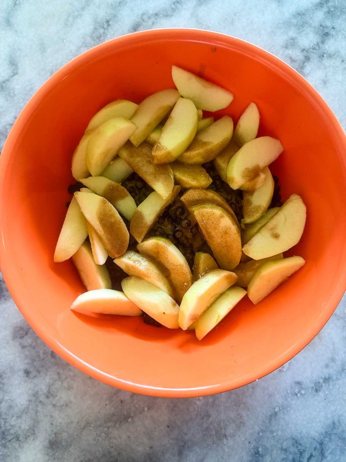 Sliced apples with cinnamon and sugar in an orange bowl, ready for mixing on a marble countertop—the perfect start for a delicious Apple Blueberry Crisp.