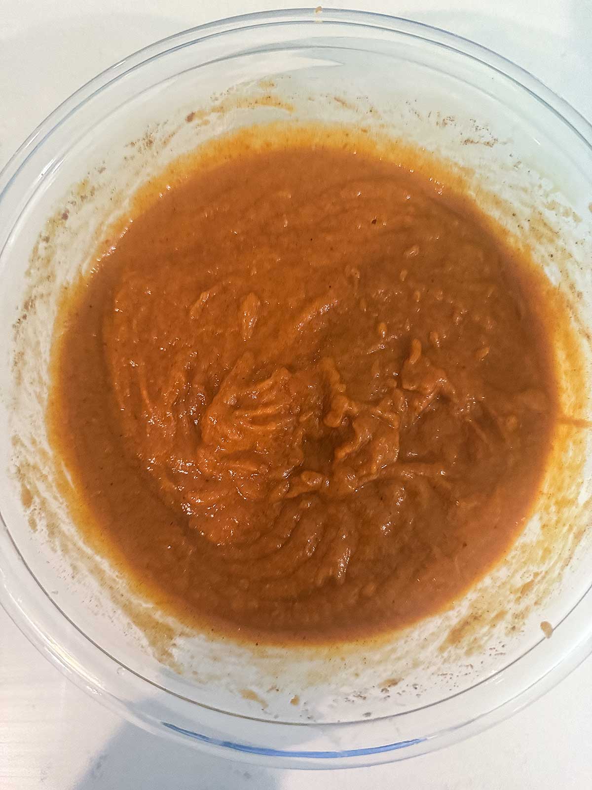 A clear bowl filled with thick, pumpkin puree in a batter, sits on a white surface. The mixture has a smooth, slightly textured consistency.