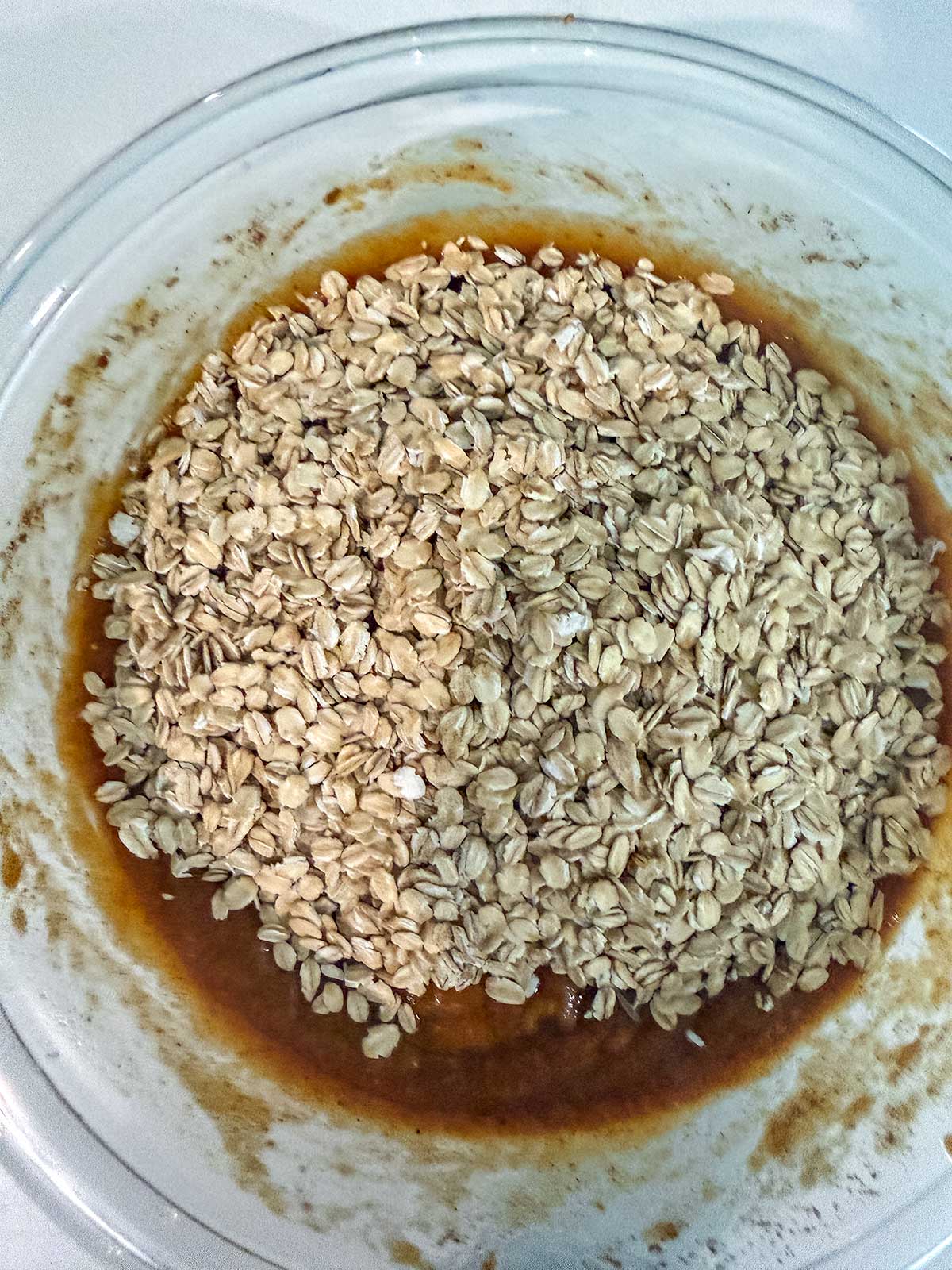 A glass bowl containing a mixture of brown batter, soon to become pumpkin oatmeal muffins, topped with a layer of dry rolled oats and ready to be mixed.