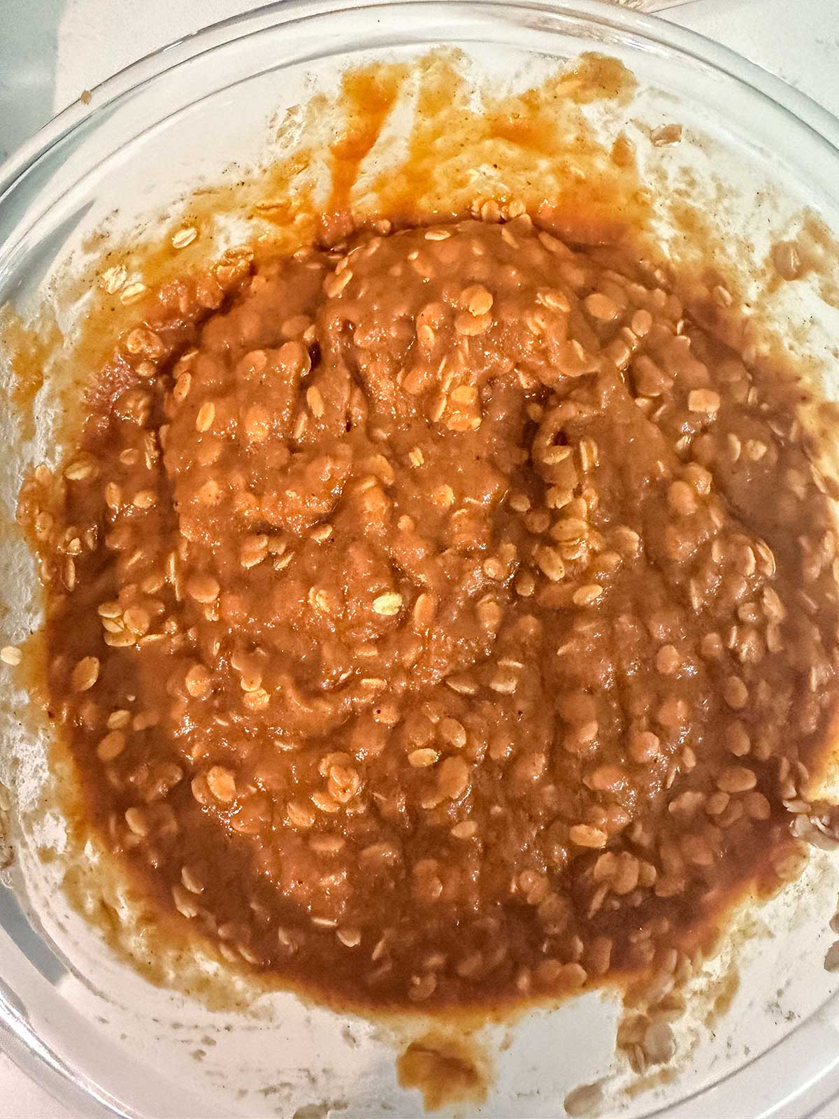 A glass bowl filled with a thick, brown mixture containing oats, likely pumpkin oatmeal muffin batter, sitting on a light-colored surface.