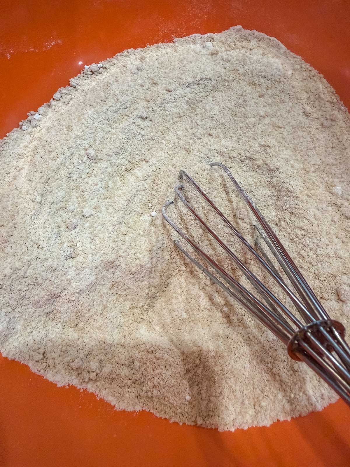 A metal whisk rests on powdered ingredients in an orange mixing bowl, suggesting the start of a pumpkin oatmeal muffins baking recipe.