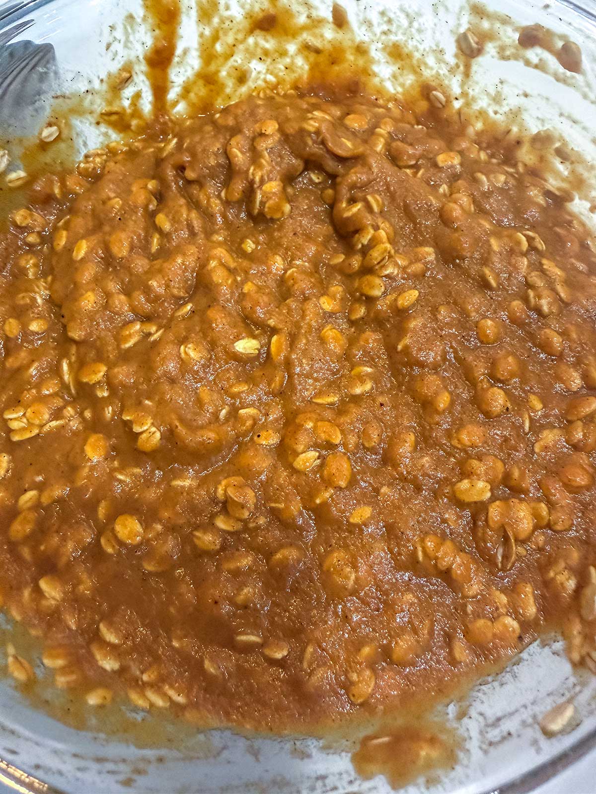 A close-up of a glass bowl filled with thick, orange-brown batter mixed with oats, likely for baking pumpkin oatmeal muffins or bars.