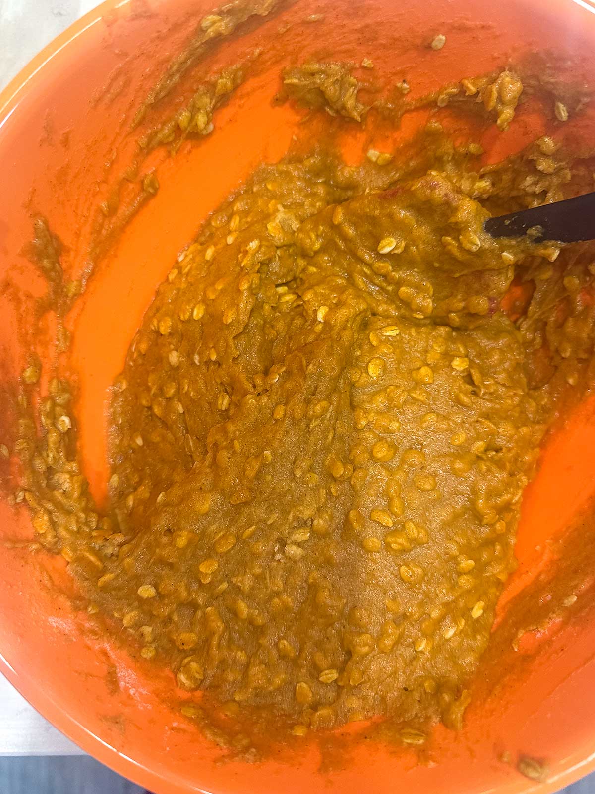 Batter for pumpkin oatmeal muffins with oats and a brown-orange color mixed in an orange bowl, with a black mixing utensil partially visible on the right side.