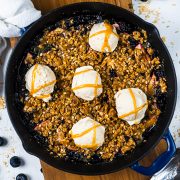 A blueberry and oat crumble, reminiscent of a classic apple blueberry crisp, is baked in a skillet, topped with five scoops of vanilla ice cream and drizzled with caramel sauce. Blueberries and oat crumbs are scattered on the table nearby.