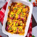 A white baking dish filled with baked chicken breasts topped with melted cheese, tomato slices, red onion, and jalapeño rings.